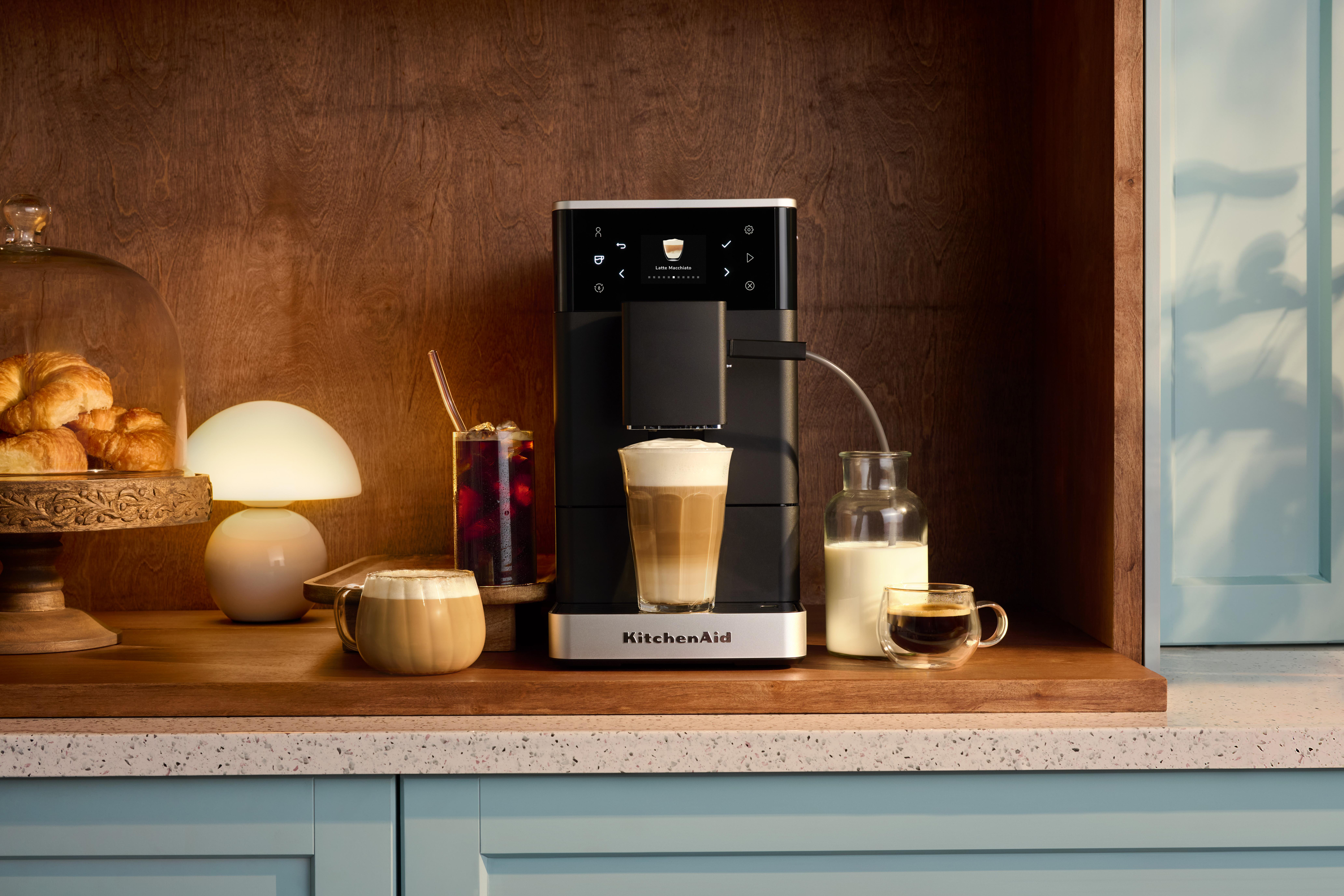 Kaffeemaschine brüht Latte Macchiato neben Croissants, Lampe und Getränken.