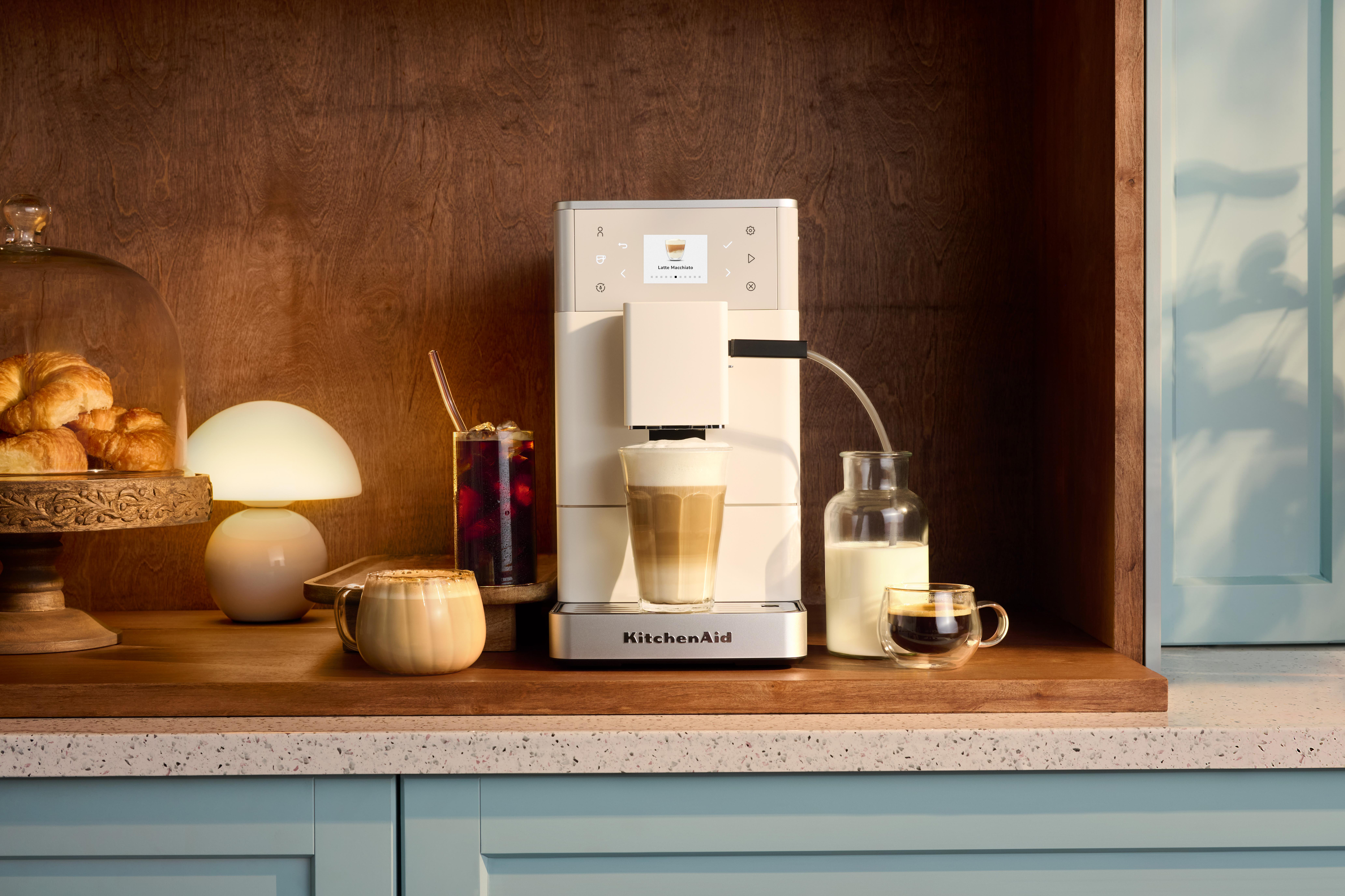 Eine weiße Kaffeemaschine macht einen Latte Macchiato, daneben stehen Kaffee und Croissants.