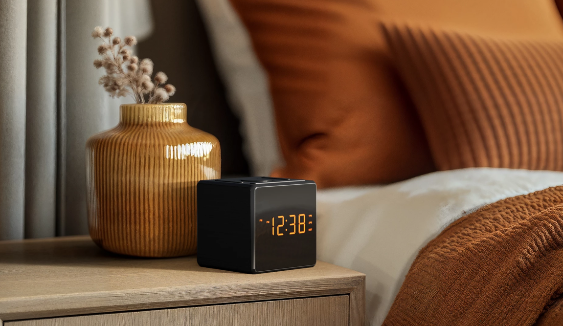 Ein schwarzer digitaler Wecker zeigt 12:38 auf einem Nachttisch neben einer Vase mit Trockenblumen.