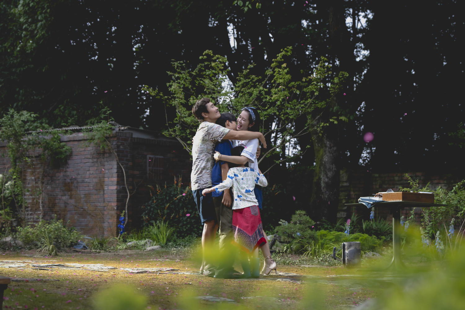 Eine Familie umarmt sich im Freien mit Bäumen und einer Ziegelmauer im Hintergrund.