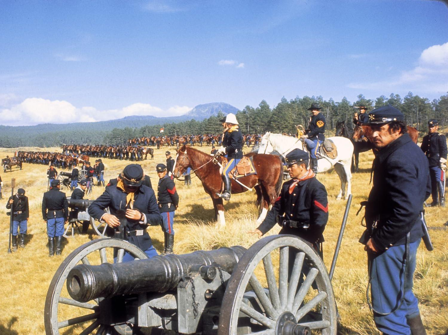 Soldaten in Uniformen mit Kanonen und Pferden in einer offenen Landschaft unter blauem Himmel.