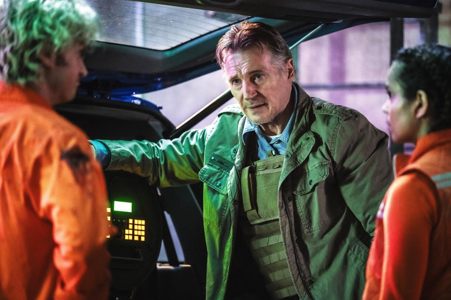 Drei Personen in einem dunklen Raum, zwei in orangefarbenen Overalls, einer in einer grünen Jacke.