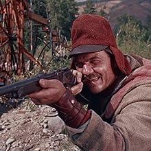 Mann in roter Mütze und Umhang zielt mit Gewehr, dahinter ein rostiges Rad.