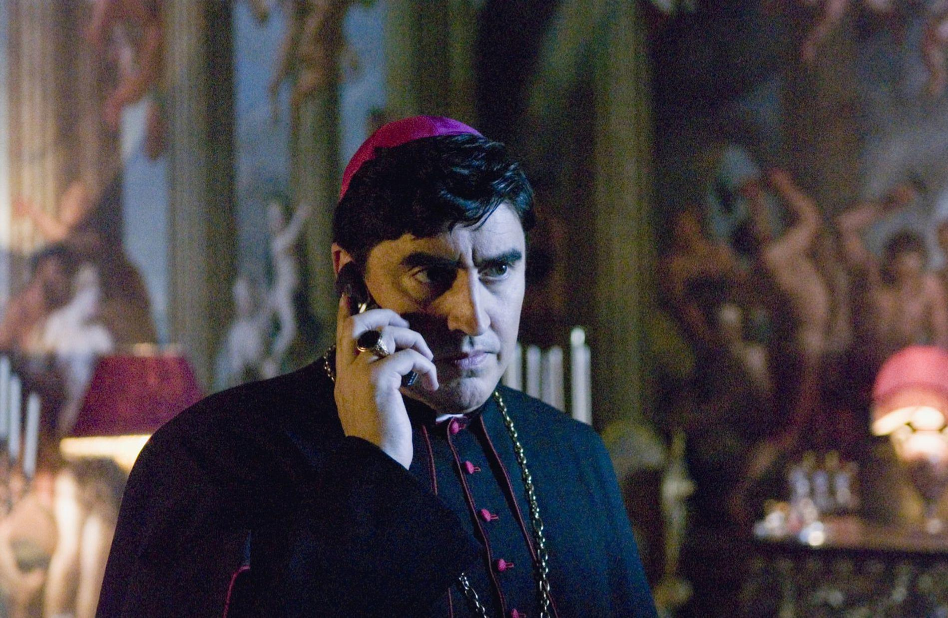 Ein Mann in bischöflicher Kleidung telefoniert vor einer bemalten Wand.