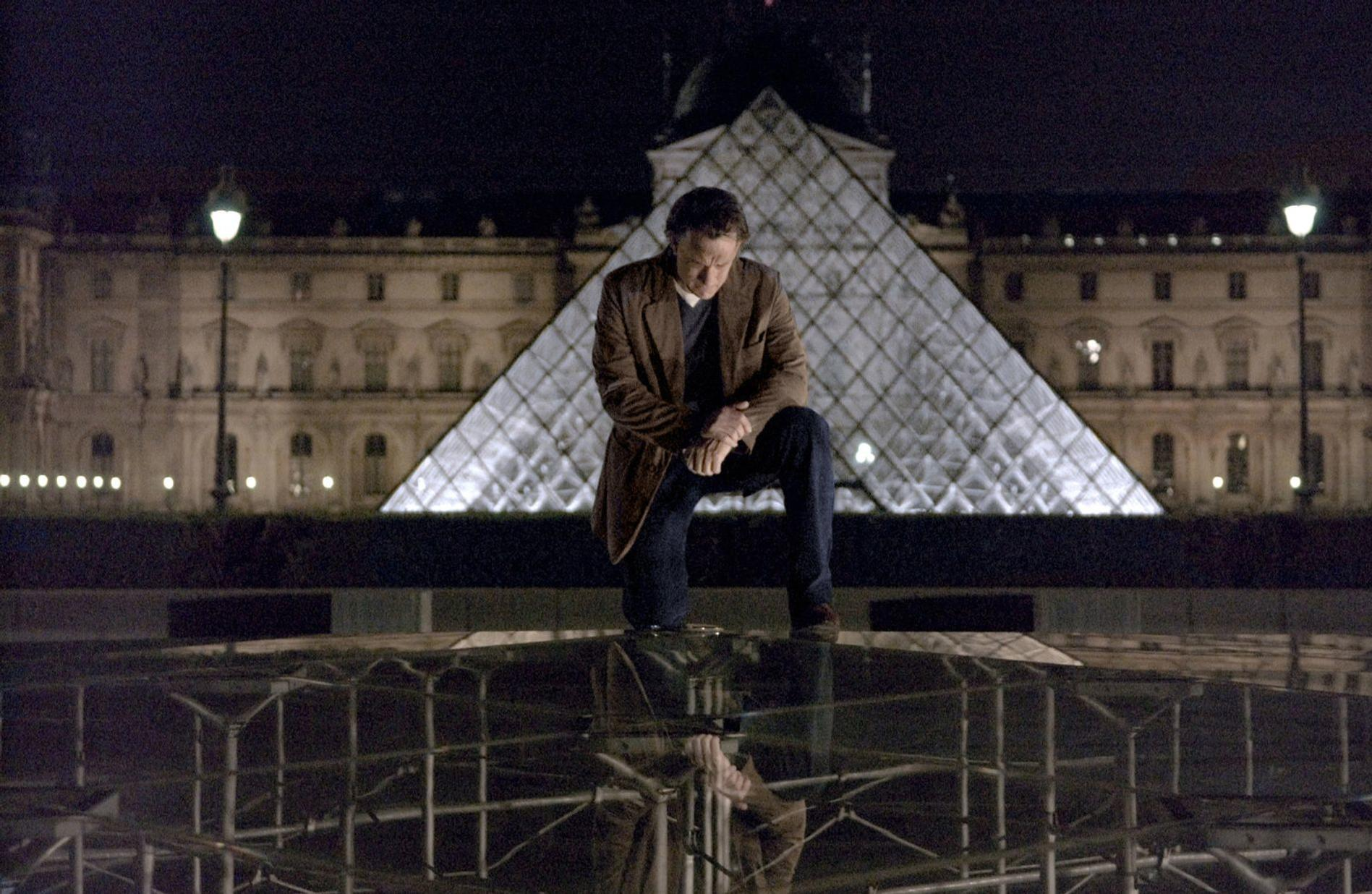 Mann kniet vor Louvre-Pyramide bei Nacht, reflektiert auf Glas.