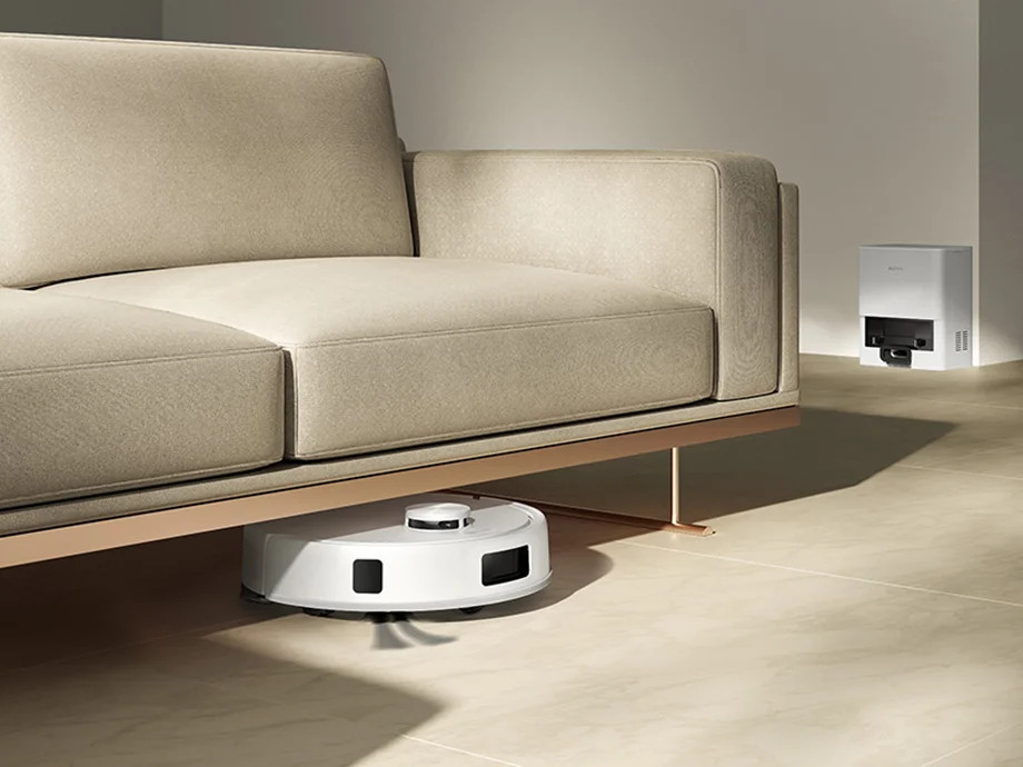 Biały robot sprzątający czyści pod beżową sofą. Stacja ładująca znajduje się na ścianie.