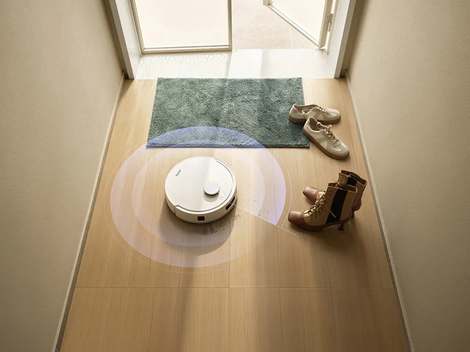 Biały robot sprzątający czyści drewnianą podłogę obok butów i dywanika.
