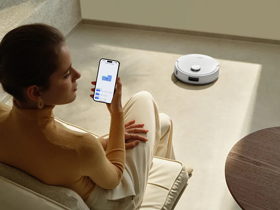 Kobieta trzyma smartfon z aplikacją, podczas gdy robot sprzątający jedzie w tle.