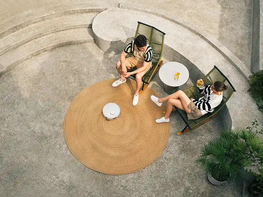 Mężczyzna i kobieta siedzą na krzesłach, robot sprzątający na dywanie.