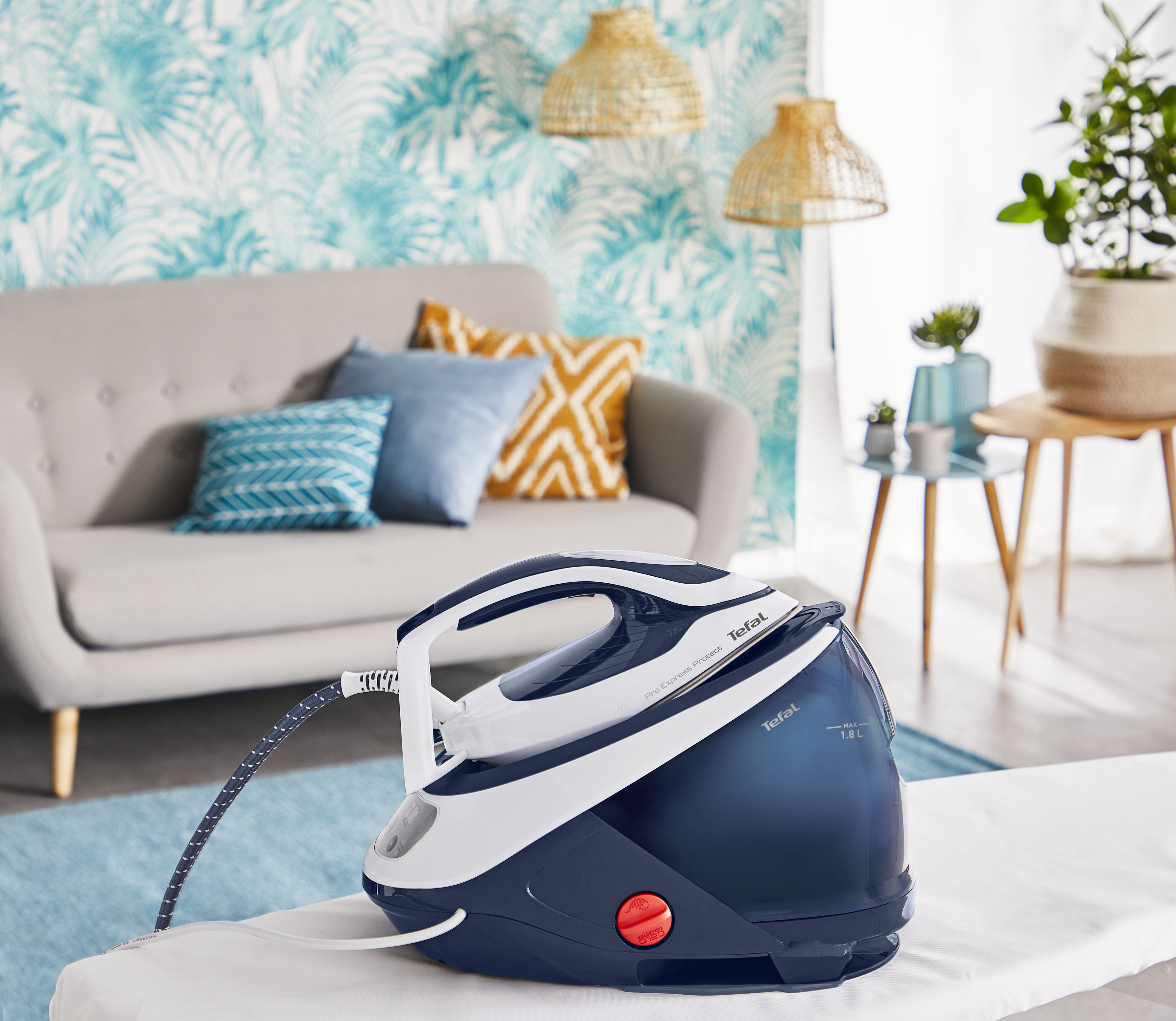 Kék és fehér Tefal gőzállomás vasaló fehér felületen, kanapé és trópusi tapéta a háttérben.