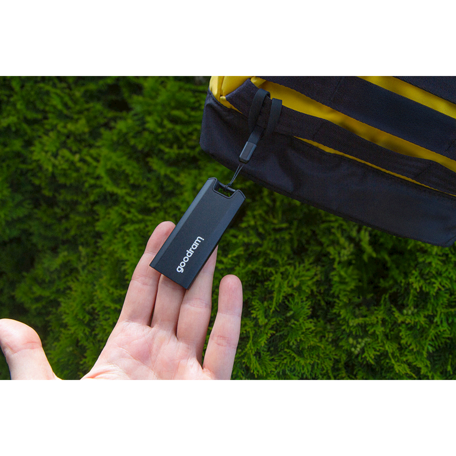 Una mano tiene una chiavetta USB nera con "goodram" davanti a una borsa e alberi.
