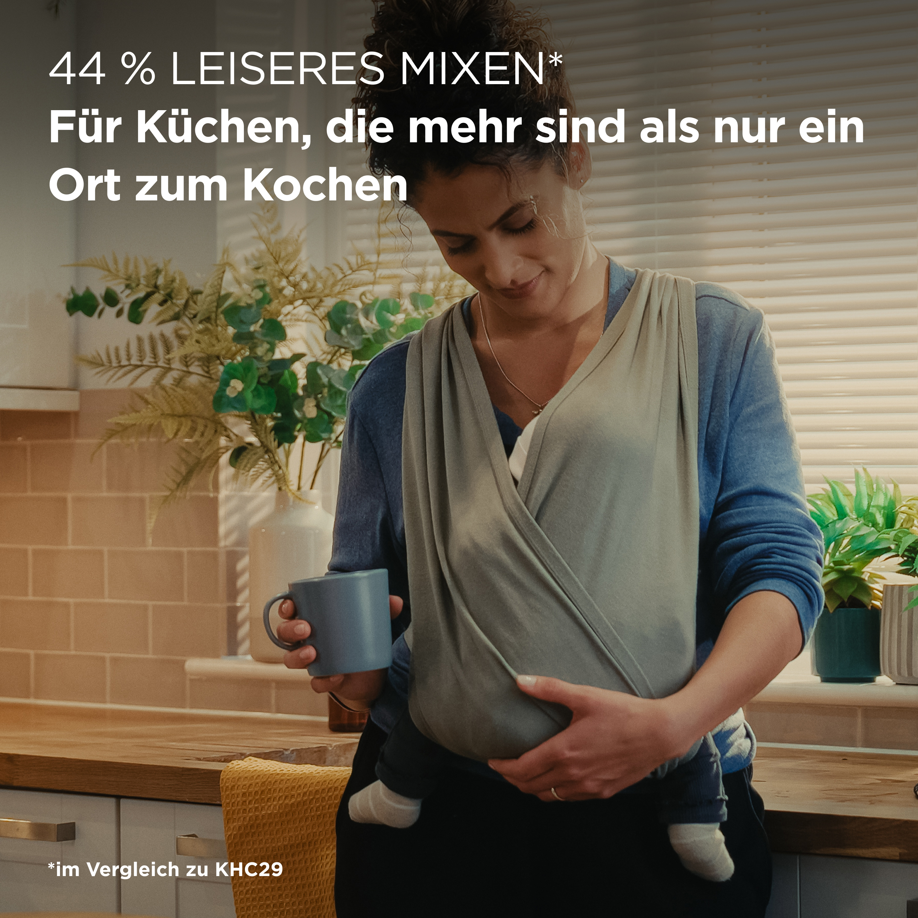 Eine Frau mit Baby im Tragetuch hält eine Tasse in einer Küche.