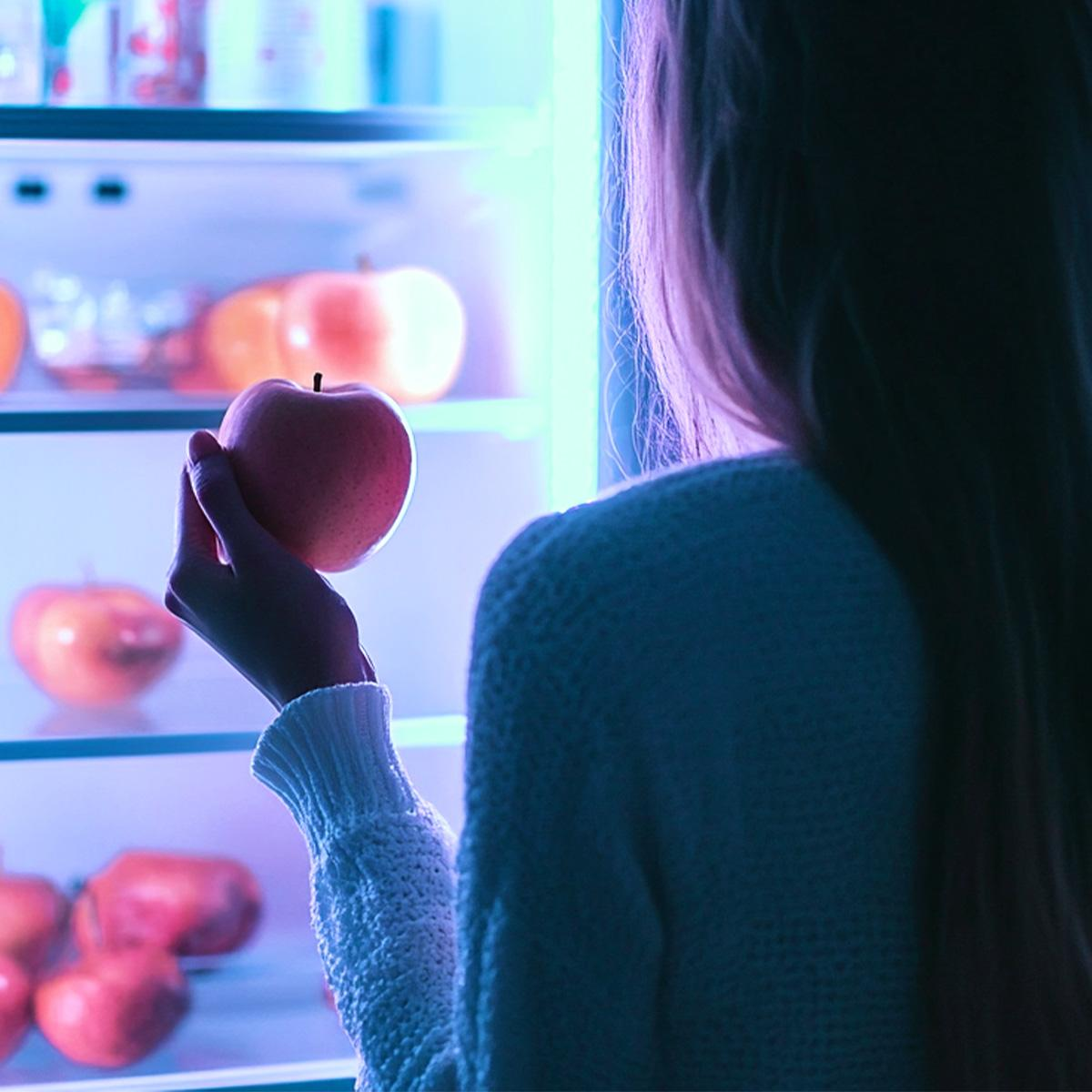 Una mujer sostiene una manzana roja frente a un refrigerador iluminado con fruta.