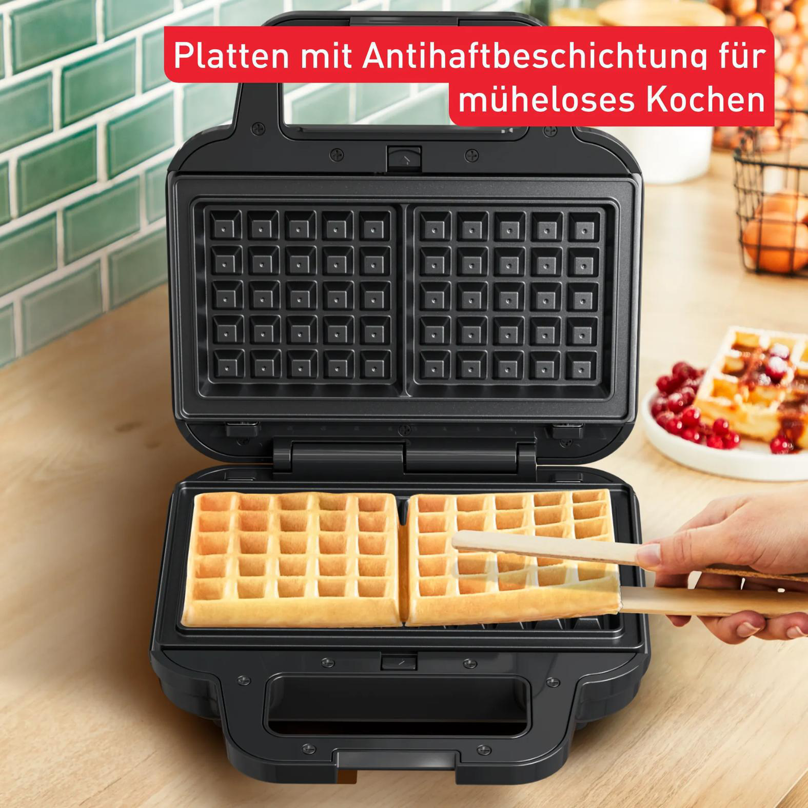 Ein offenes Waffeleisen mit zwei goldbraunen Waffeln, eine wird mit Holzstäbchen herausgenommen.