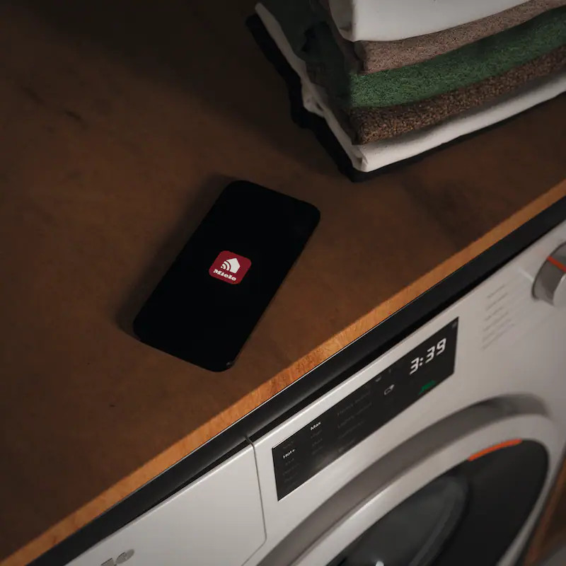 Smartfon z aplikacją Miele leży na drewnianej szafce obok pralki.