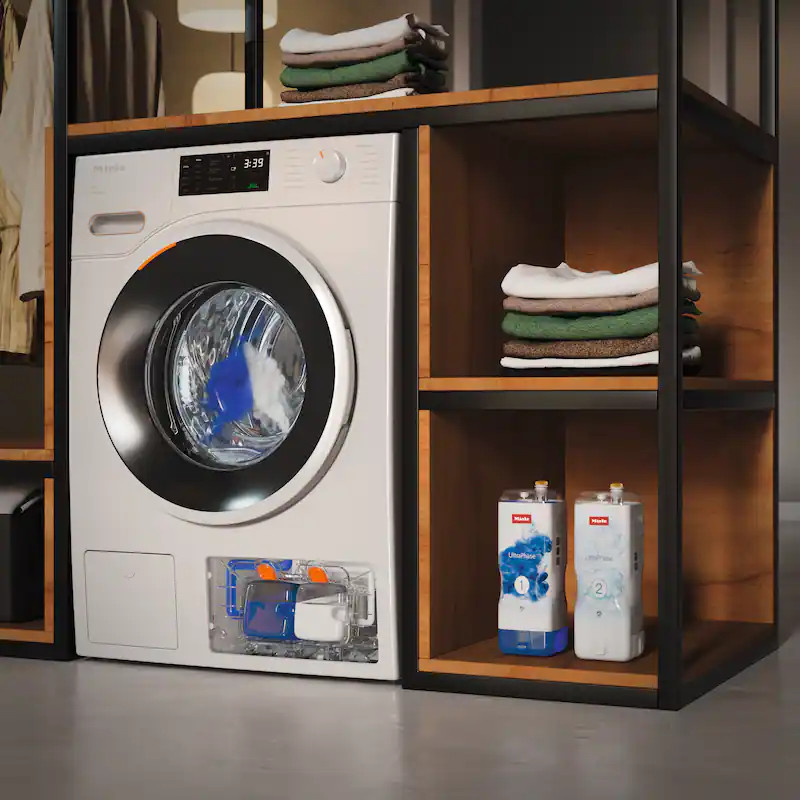 Biała pralka Miele z przezroczystym dozownikiem na detergent i stosami ręczników na półkach.