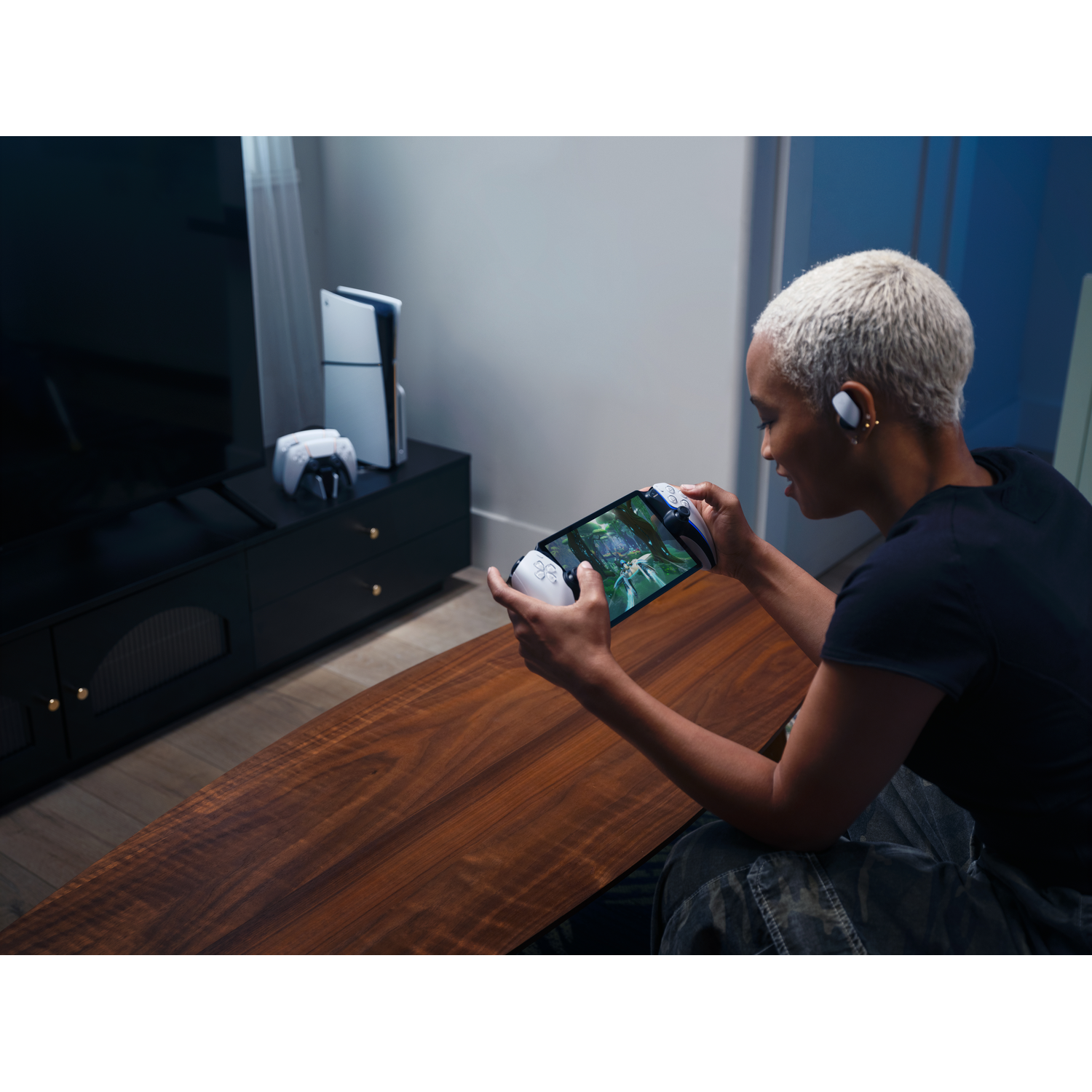 A person plays a video game on a handheld device, with a console and controllers in the background.