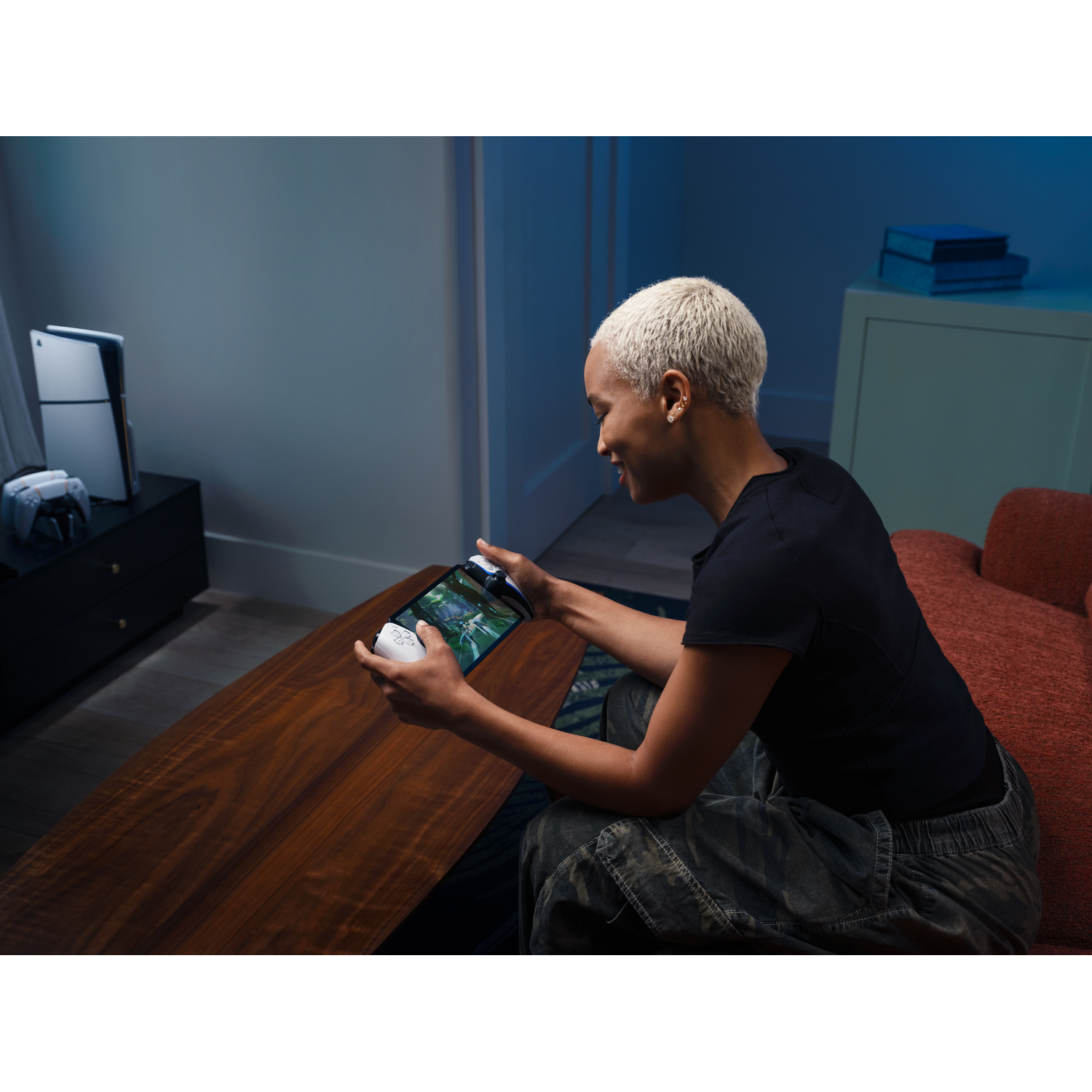 A person plays on a handheld device, with a game console in the background.