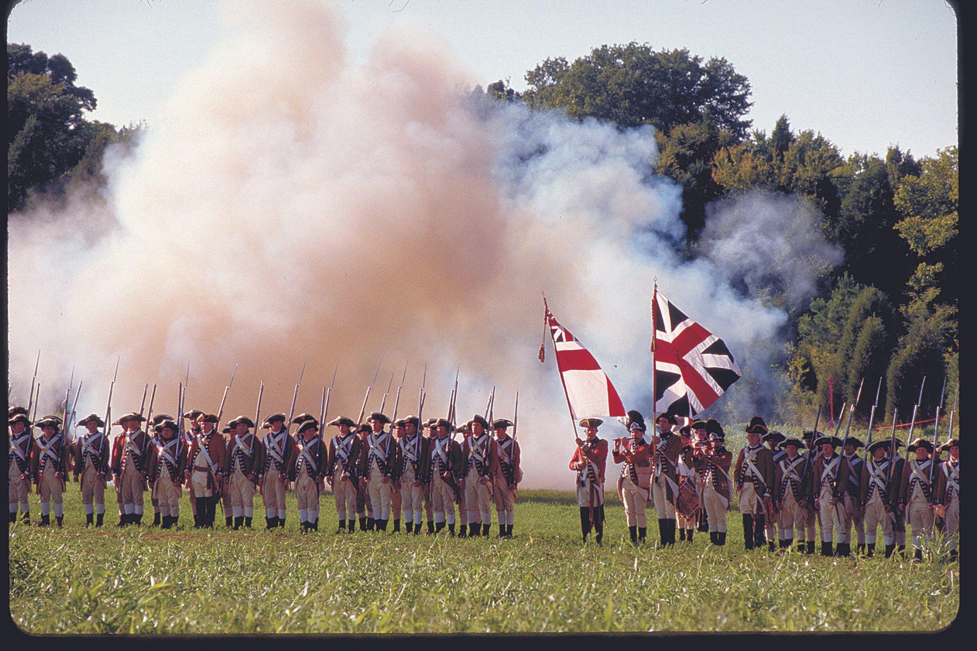 Soldaten in historischen Uniformen stehen in einer Reihe, Fahnen und Rauch im Hintergrund.