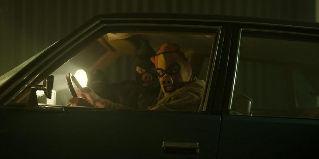Zwei Personen in Masken sitzen in einem Auto. Einer fährt, der andere blickt nach rechts.