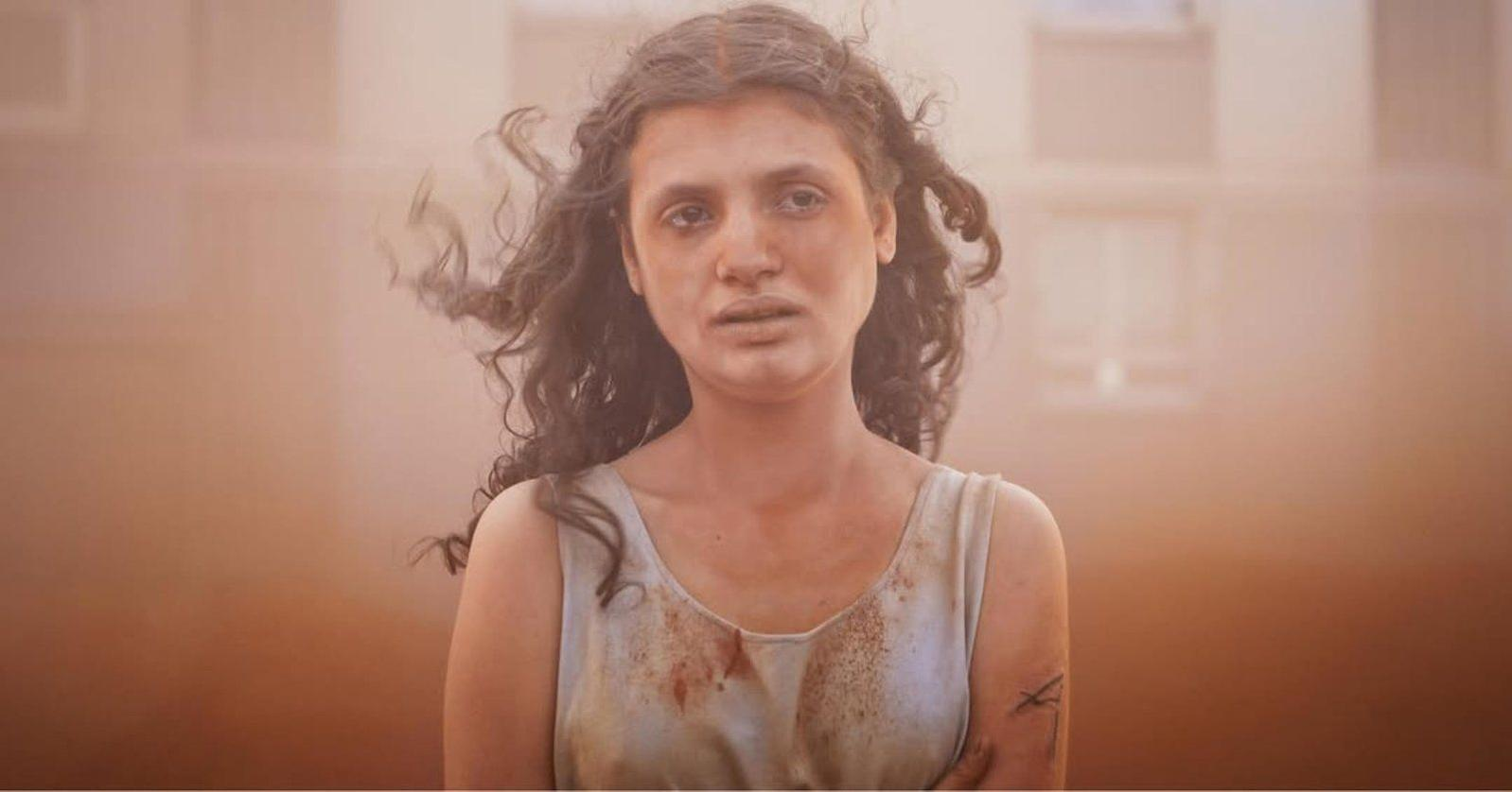 Eine Frau mit dunklen, lockigen Haaren trägt ein schmutziges, helles Kleid. Der Hintergrund ist unscharf und orangefarben.