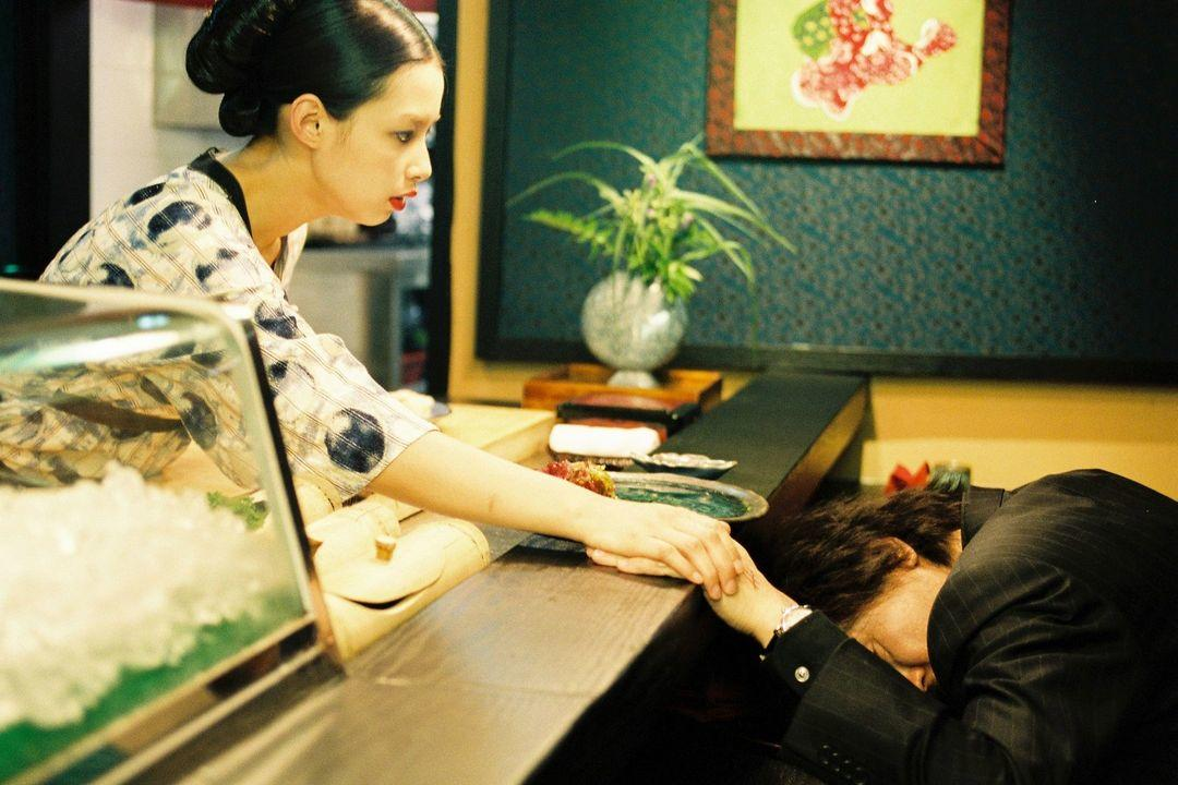 Eine Frau im Kimono reicht einem Mann, der seinen Kopf auf den Tisch gelegt hat, die Hand.
