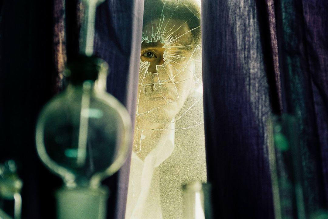 Ein Auge blickt durch ein zerbrochenes Fenster, umgeben von lila Vorhängen.