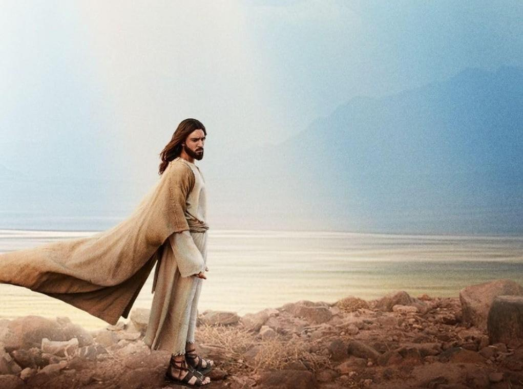 Jesus mit braunem Gewand und Tuch, steht auf felsigem Ufer am Wasser.