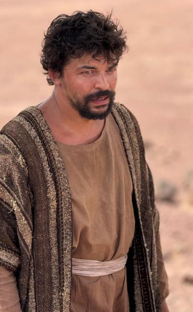 Ein Mann mit dunklem, lockigem Haar und Bart trägt eine braune Tunika und eine verzierte Robe.