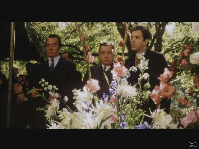 Männer in Anzügen stehen hinter Blumen bei einer Trauerfeier im Freien mit Bäumen im Hintergrund.