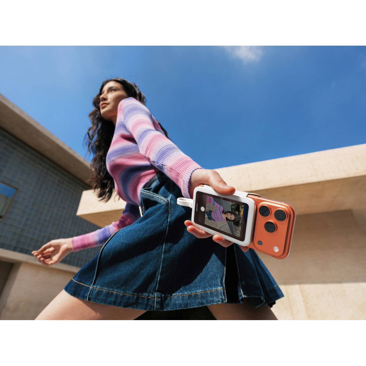 Una donna dai capelli scuri indossa un top a righe e una gonna di jeans, tenendo uno smartphone arancione.