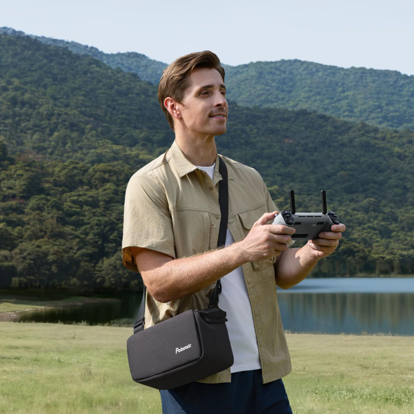 Mann mit Fernbedienung und Tasche steht im Freien vor Hügeln und See.