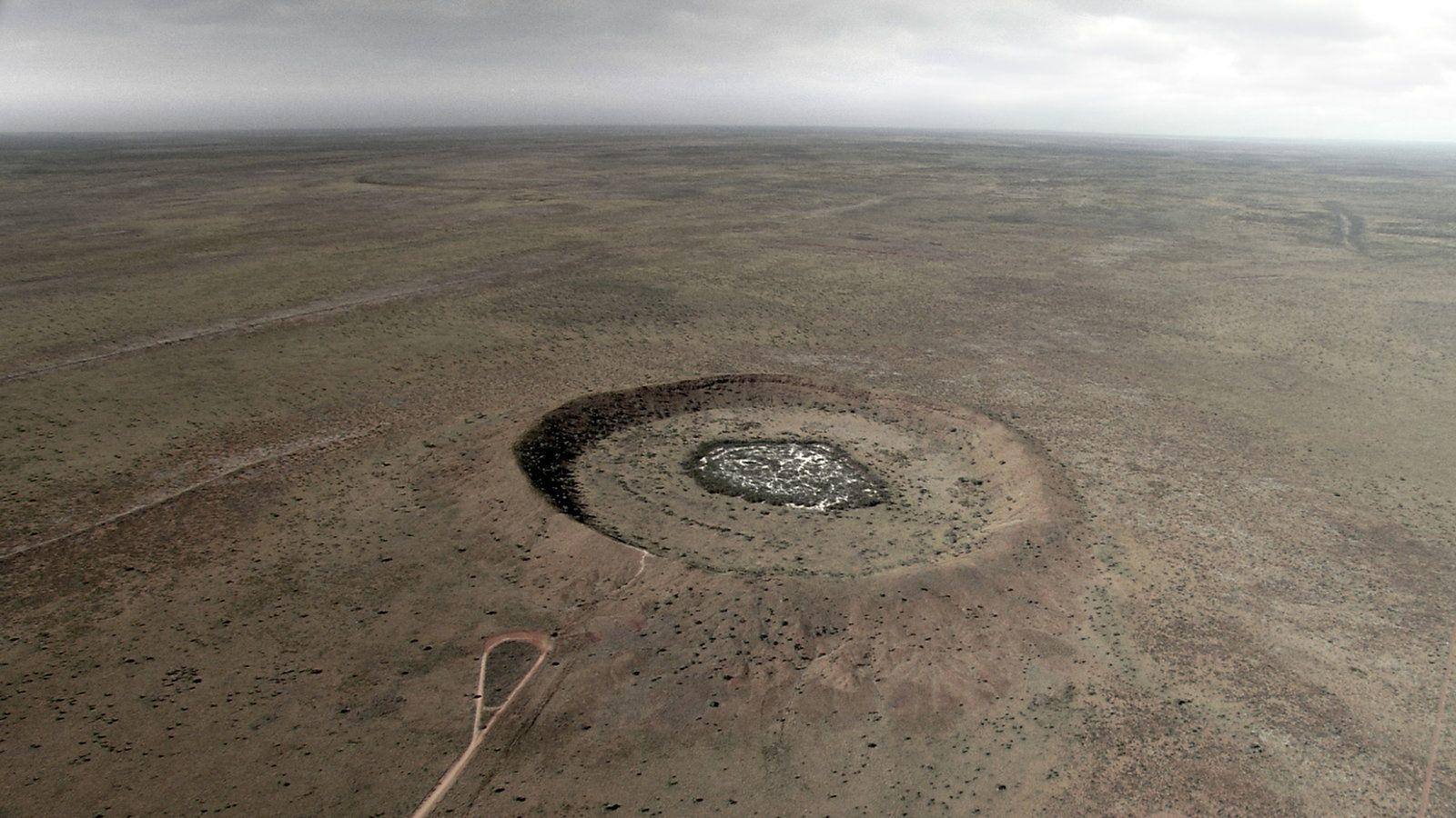 Eine riesige, kreisförmige Kraterformation in einer flachen, trockenen Landschaft unter einem bewölkten Himmel.