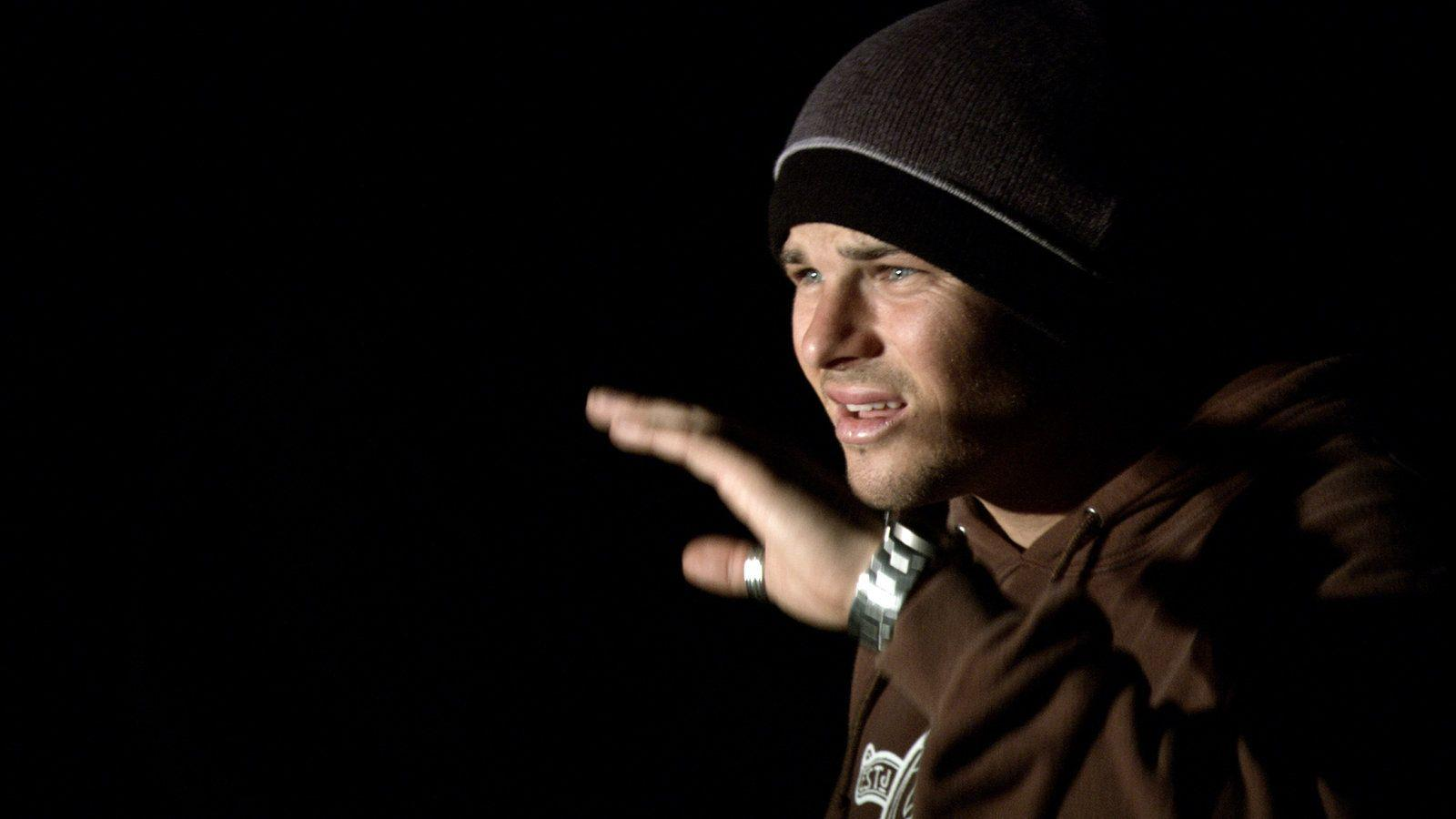 Ein Mann mit Beanie und Kapuzenpullover mit erhobener Hand, vor dunklem Hintergrund.