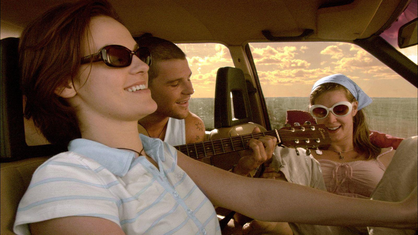 Drei Freunde reisen im Auto, einer spielt Gitarre, bei Sonnenuntergang
