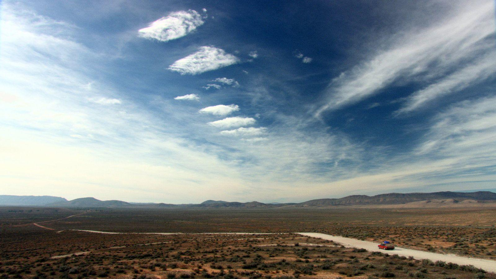 Ein rotes Auto fährt auf einer Schotterstraße durch eine weite, trockene Landschaft unter einem dramatischen Himmel.