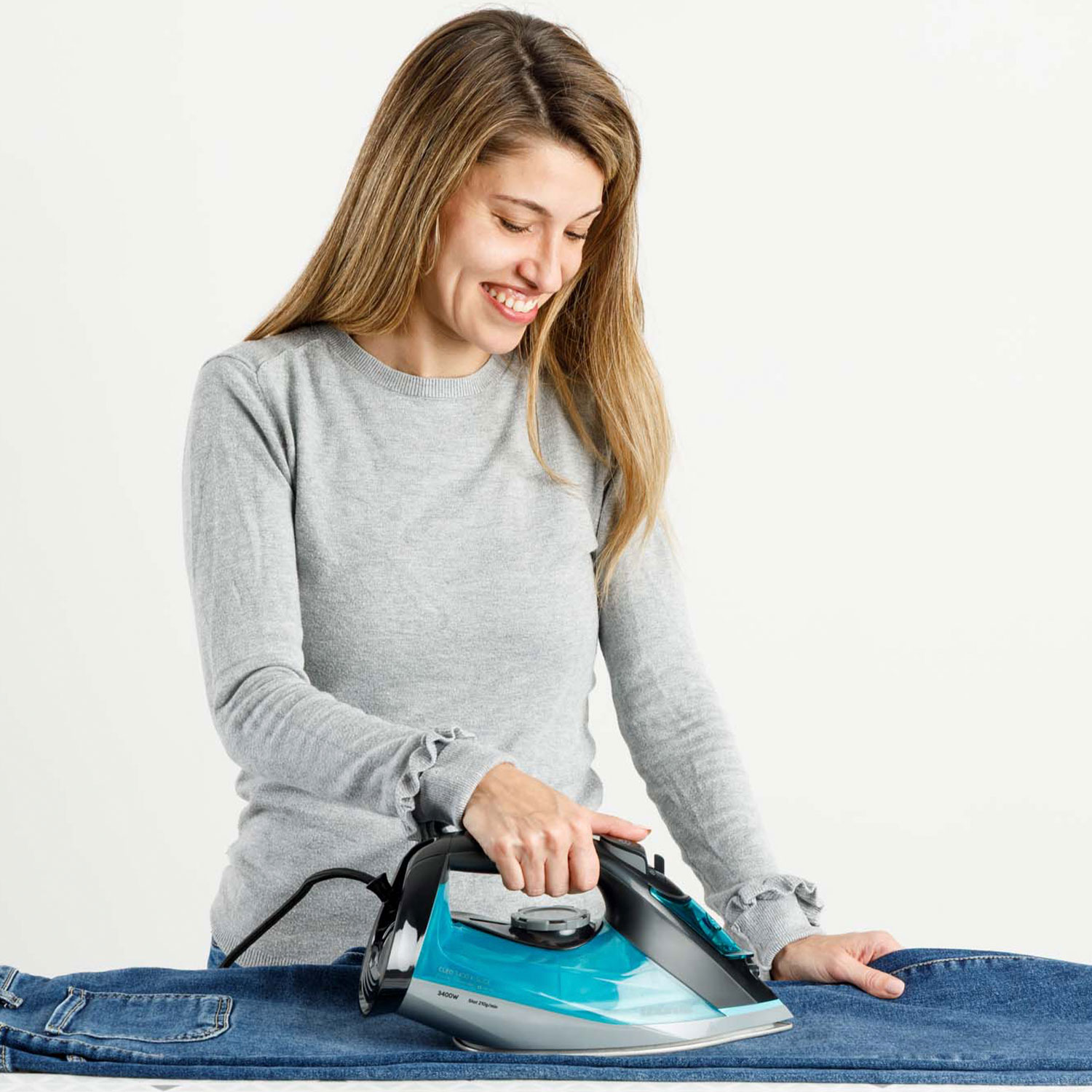 Una mujer plancha vaqueros azules con una plancha turquesa sobre fondo blanco.