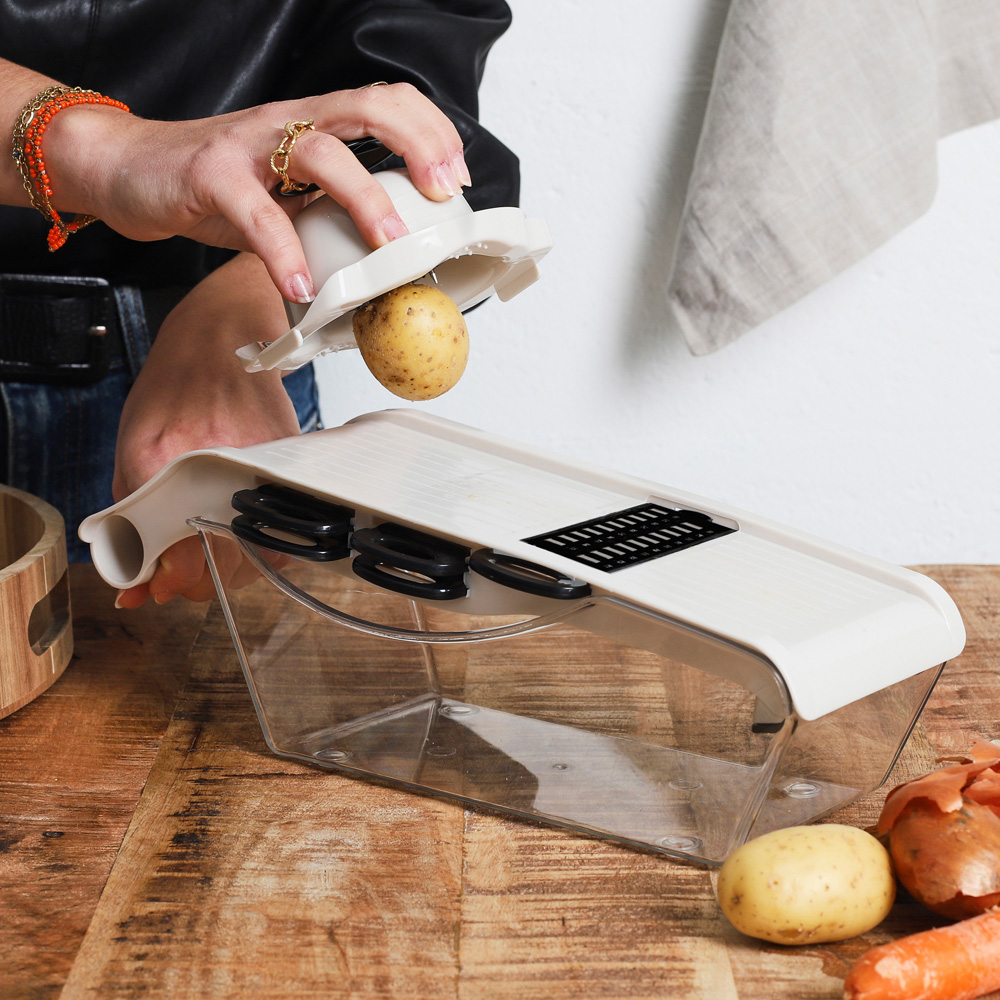 Una mano sujeta una patata sobre un cortador de verduras en una mesa de madera.