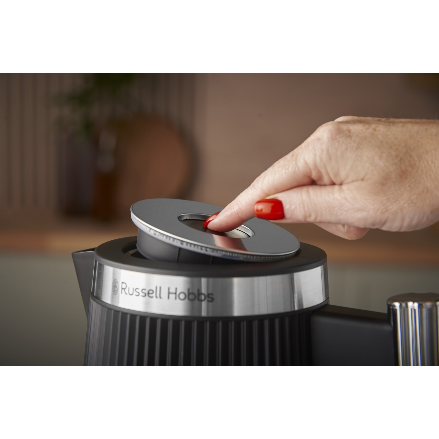 Een hand met rode nagels drukt op het deksel van een zwarte Russell Hobbs waterkoker.
