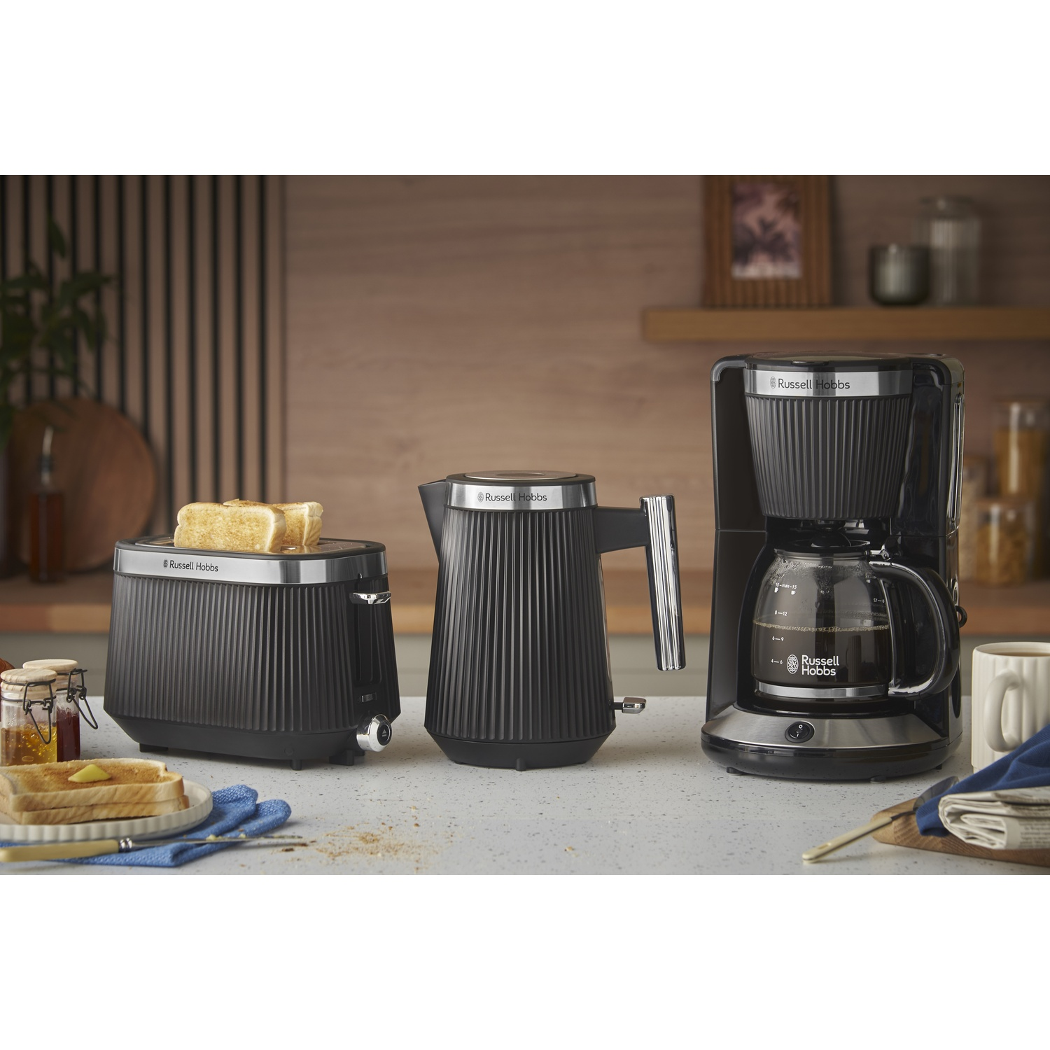 Grille-pain, bouilloire et cafetière noirs sur un plan de travail avec toasts et confiture.
