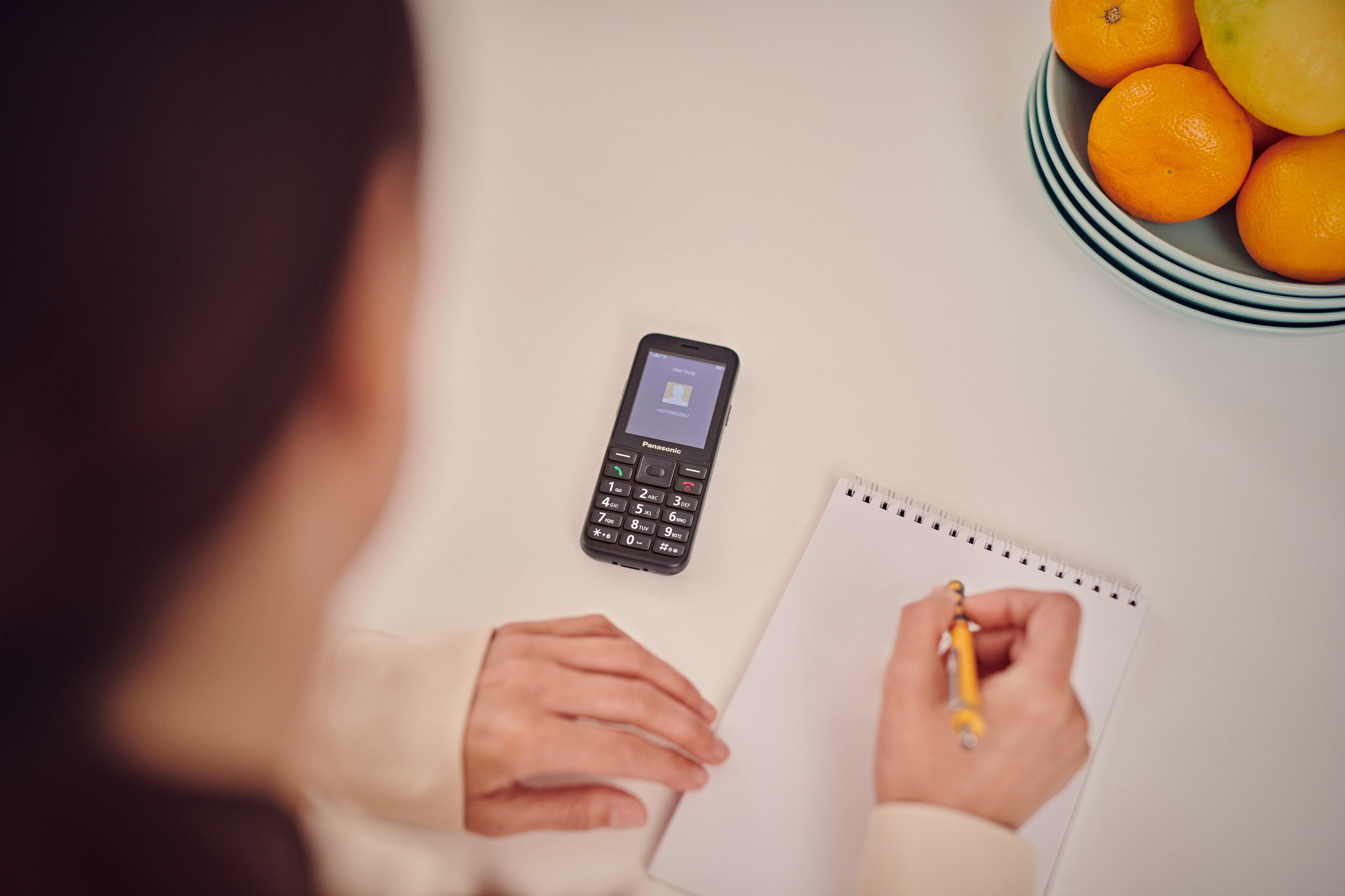 Eine Person schreibt in ein Notizbuch, daneben liegt ein Handy und eine Obstschale.