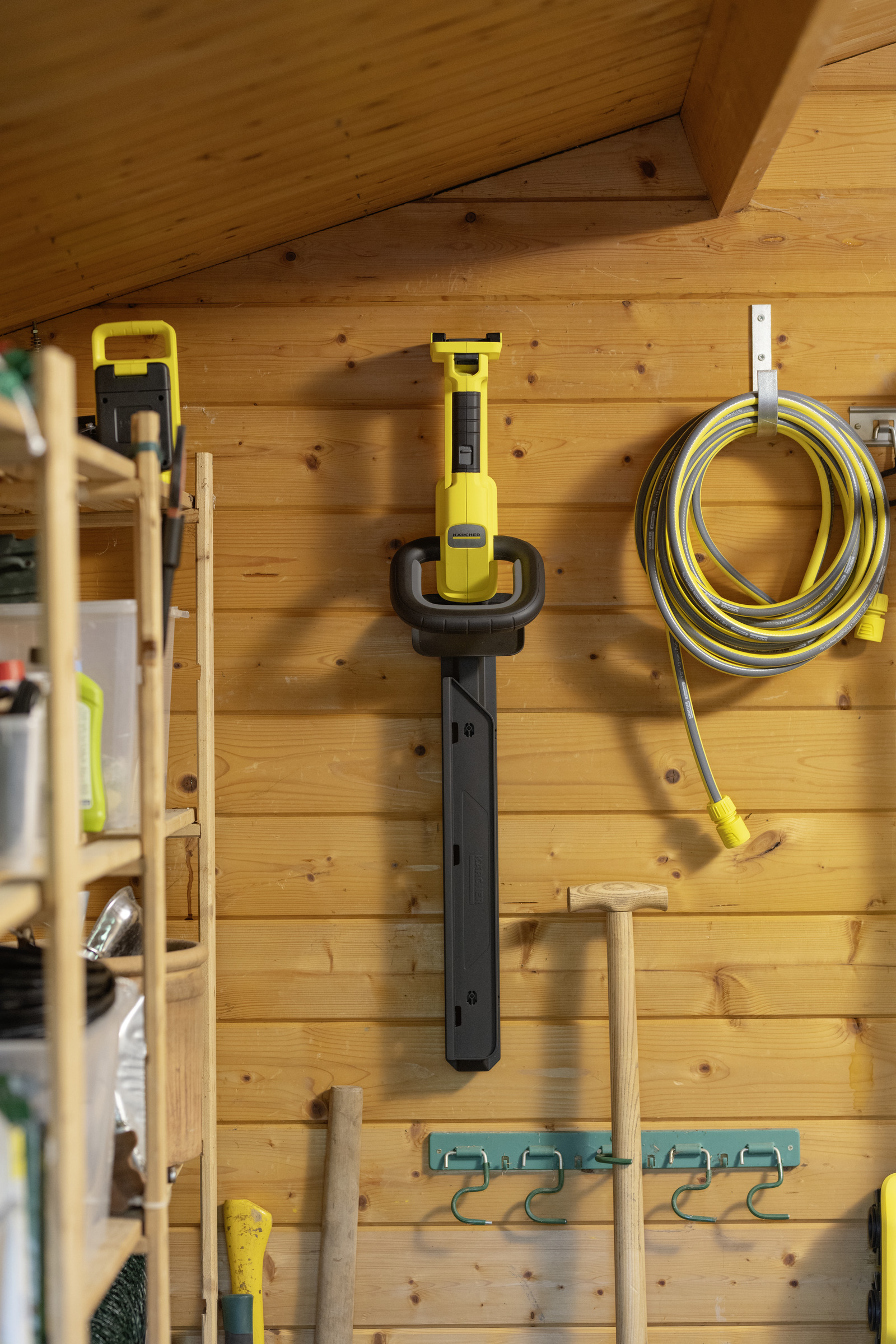 Żółta kosiarka do żywopłotu wisi na drewnianej ścianie obok węża.