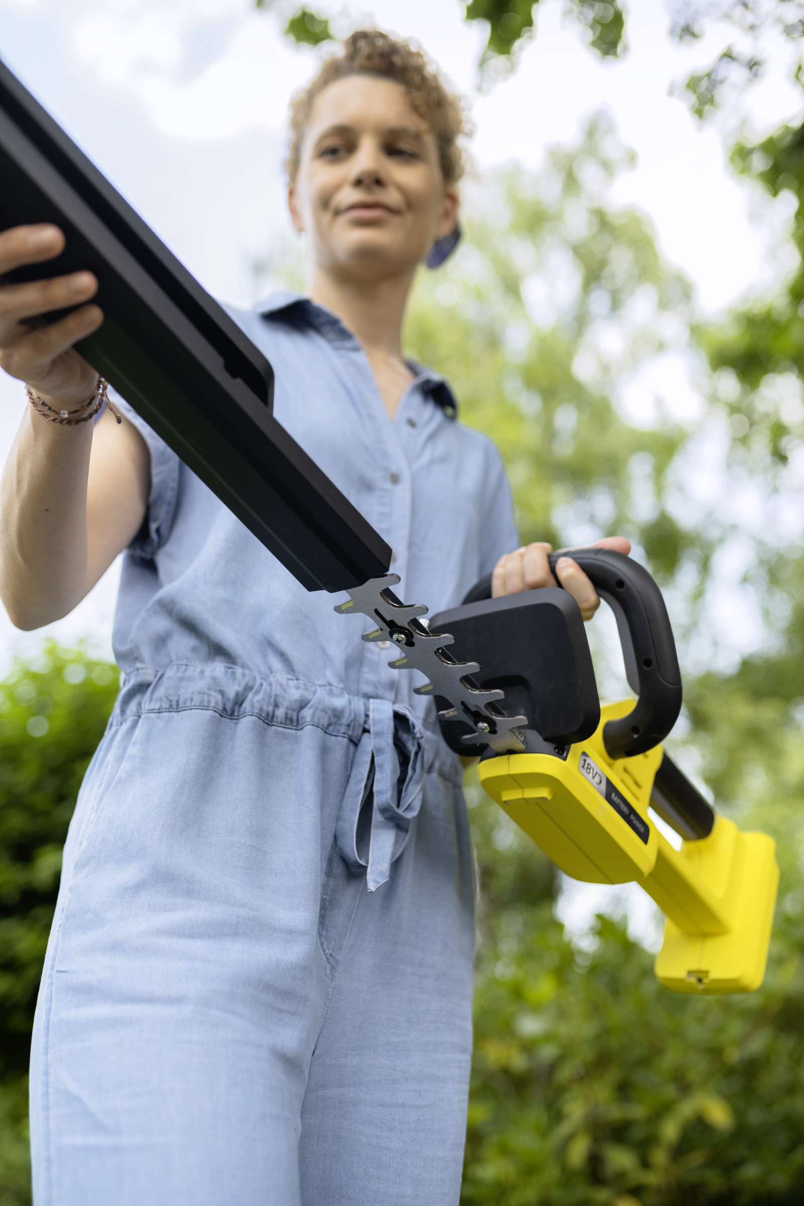 Kobieta w niebieskim kombinezonie trzyma żółtą przycinarkę do żywopłotu. Zielone drzewa w tle.