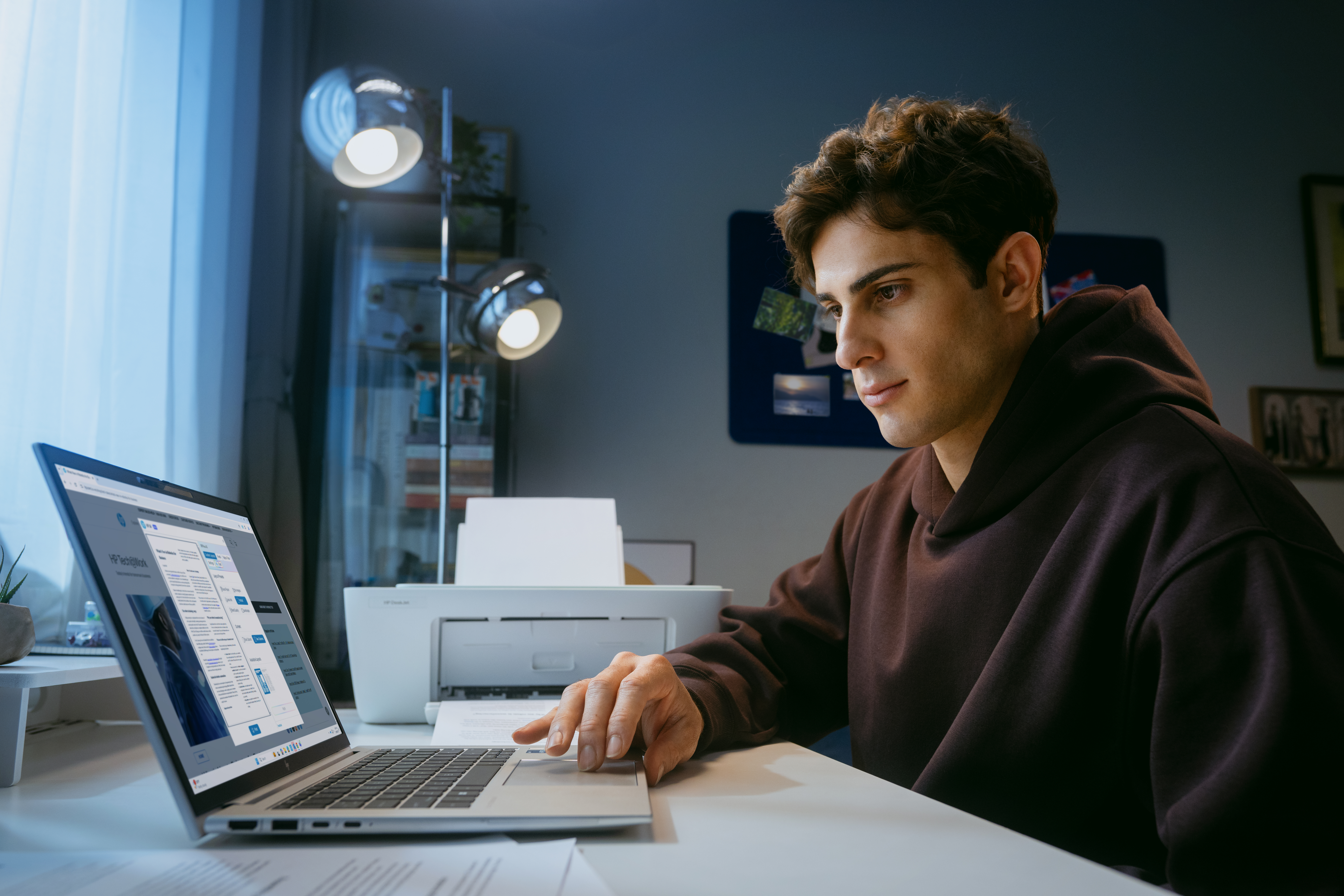 Egy férfi íróasztalnál ül laptopot használva. Egy nyomtató mellette van.