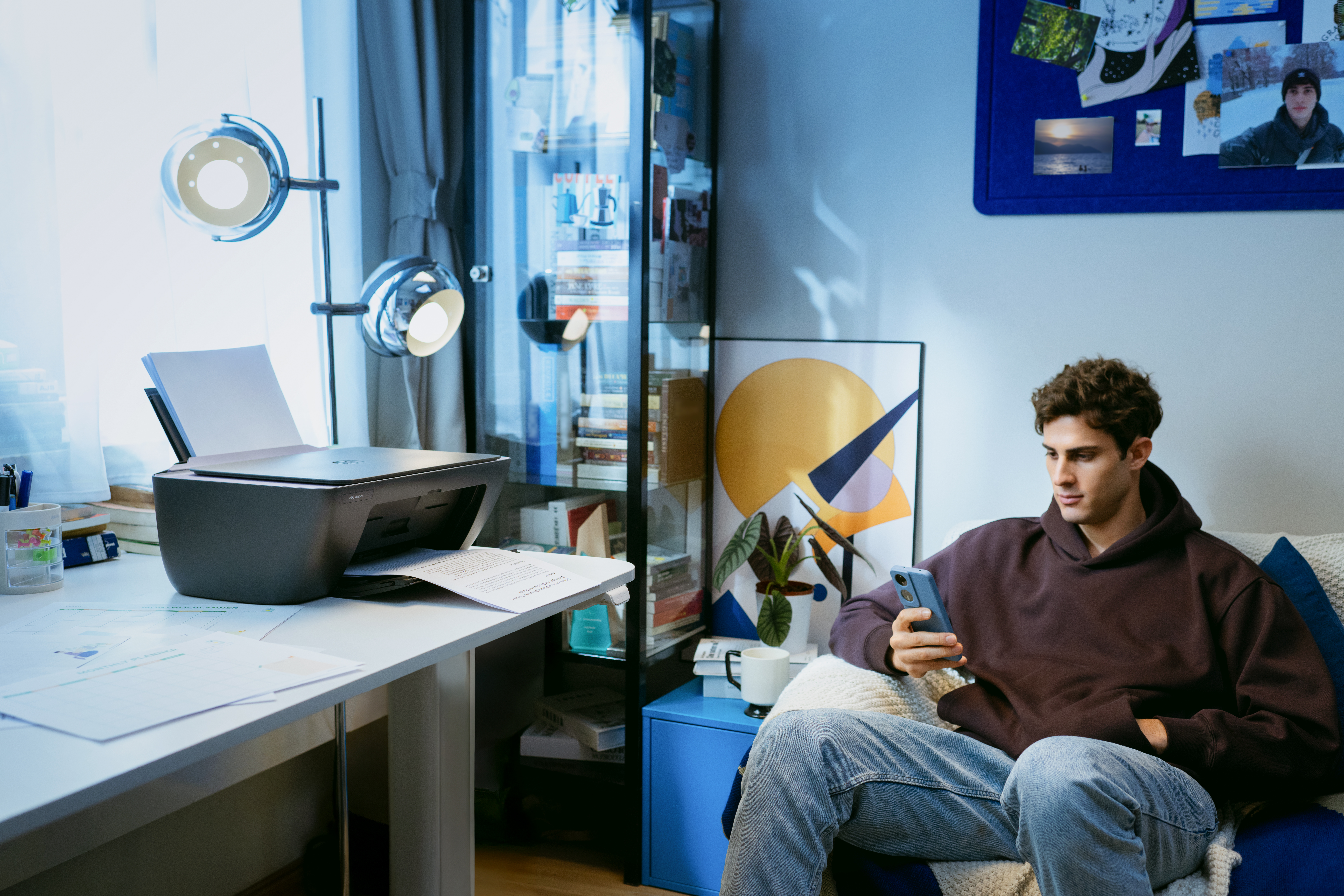 Ein Mann sitzt auf einer Couch und benutzt sein Handy, ein Drucker steht auf einem Schreibtisch.