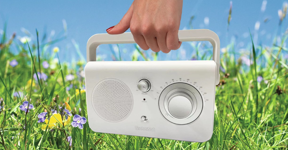 Ręka trzyma białe radio z uchwytem przed trawiastym polem z kwiatami i błękitnym niebem.