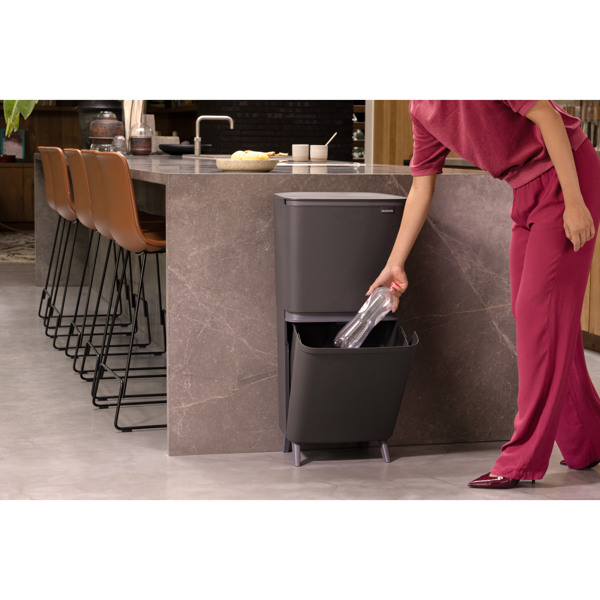 Eine Person wirft eine Plastikflasche in einen dunklen Mülleimer in einer modernen Küche.