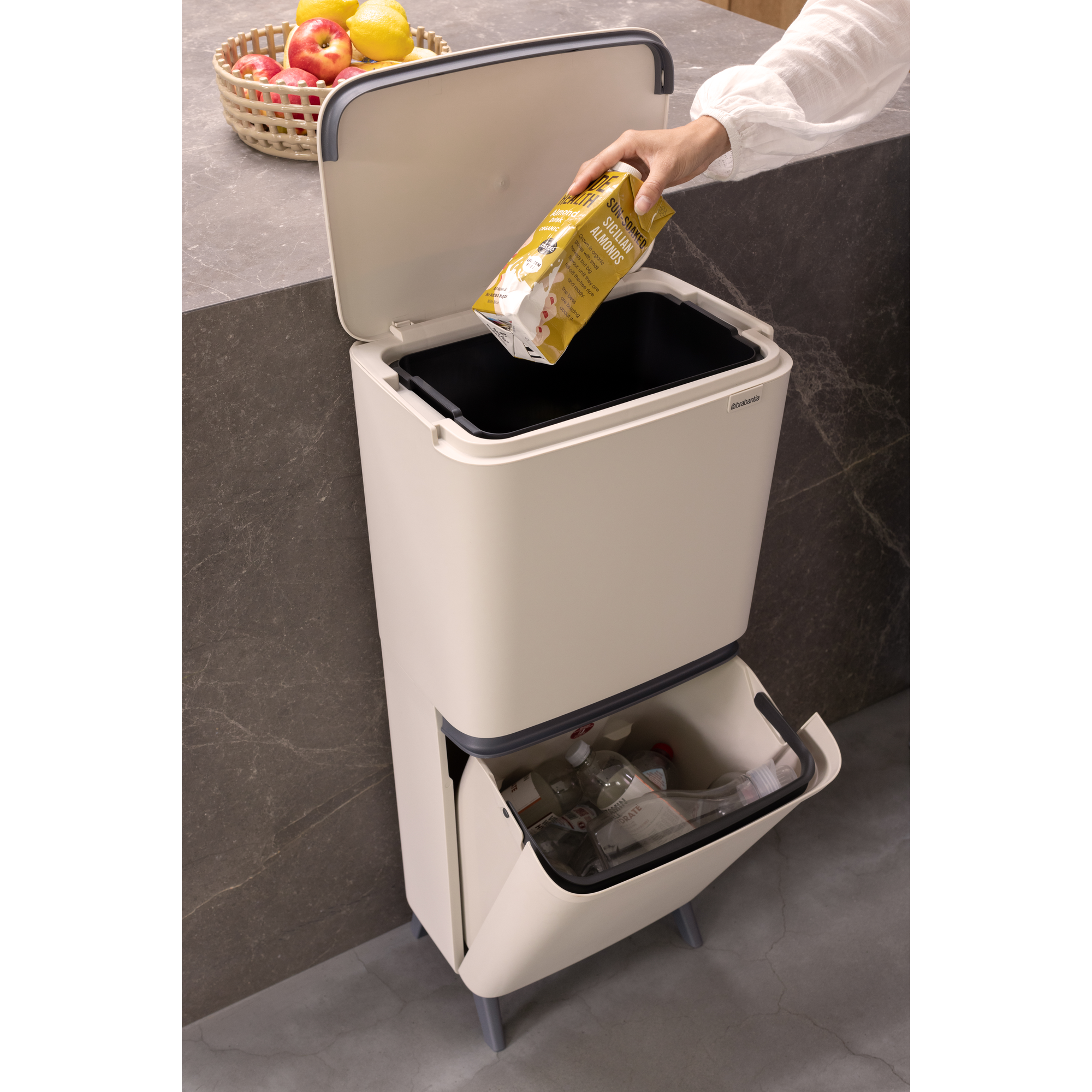 Eine Hand wirft einen Karton in einen beigen Brabantia-Recyclingbehälter. Ein Obstkorb steht daneben.