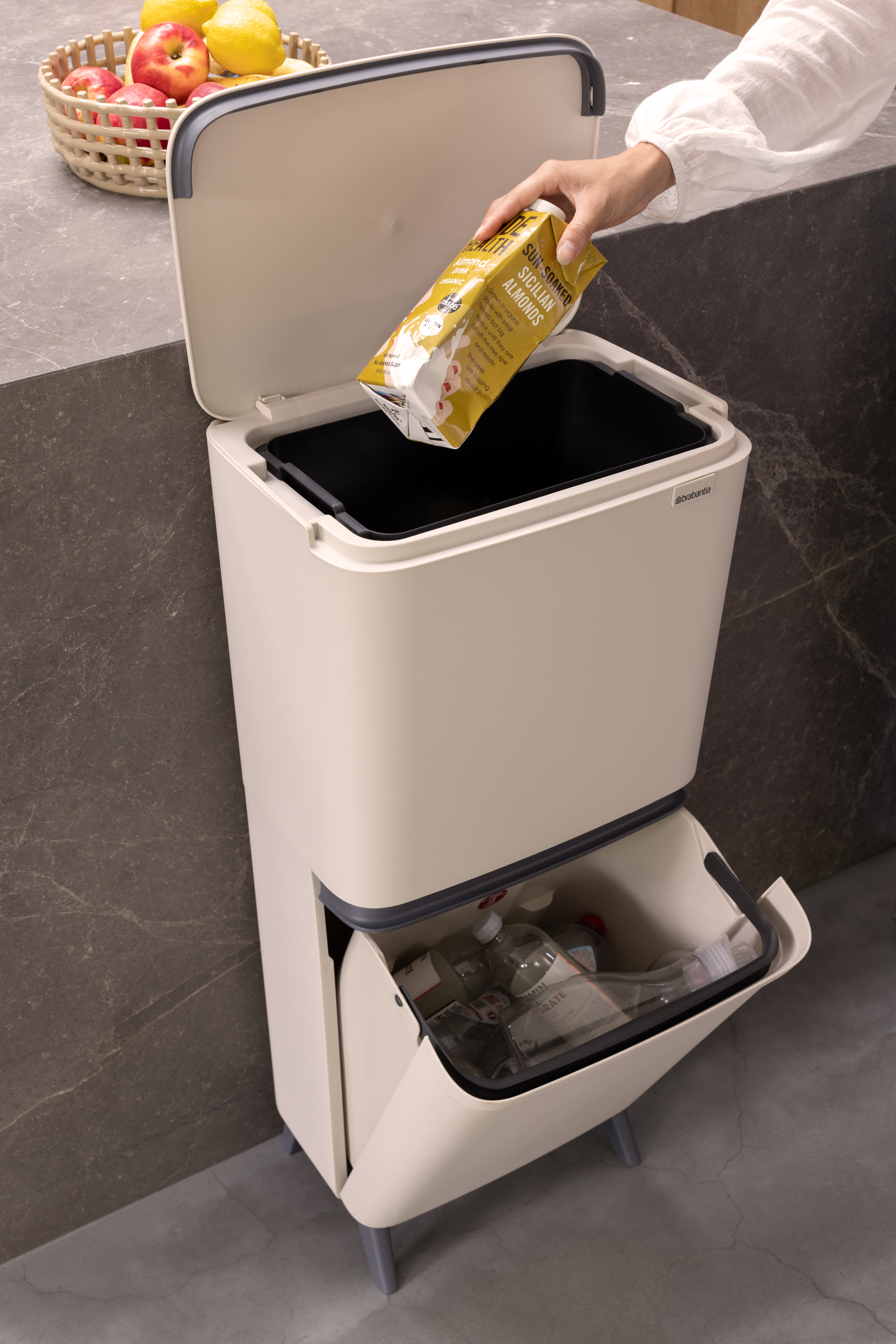 Eine Hand wirft einen Mandeldrink-Karton in einen beigen Recyclingbehälter mit zwei Fächern.
