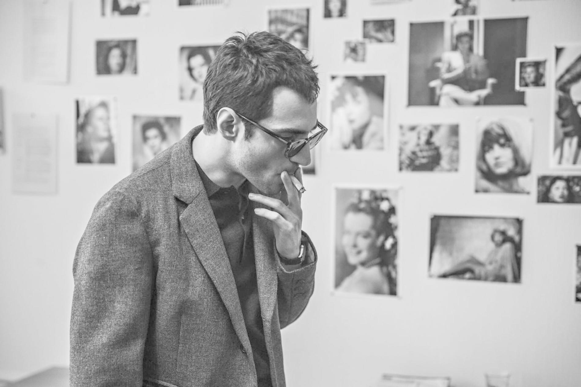 Ein Mann mit Brille raucht eine Zigarette vor einer Wand voller Schwarz-Weiß-Fotos.